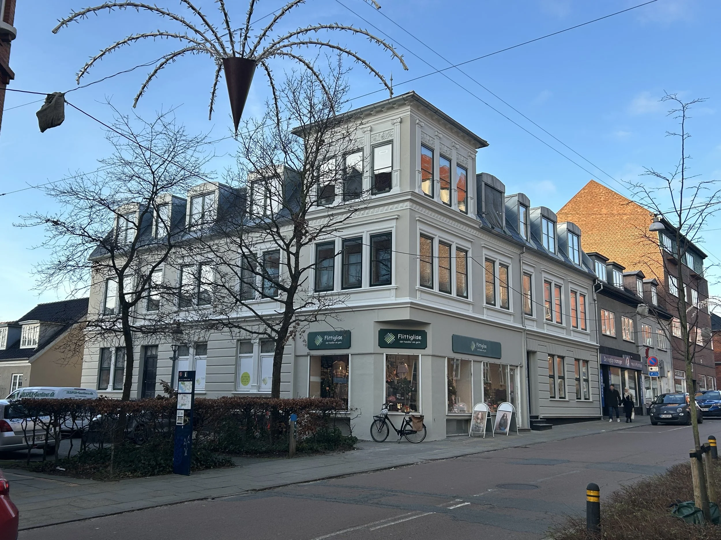 En hvid, tre-etagers bygning ved en gadehjørne med flere vinduer og tagvinduer, omgivet af træer og buske.