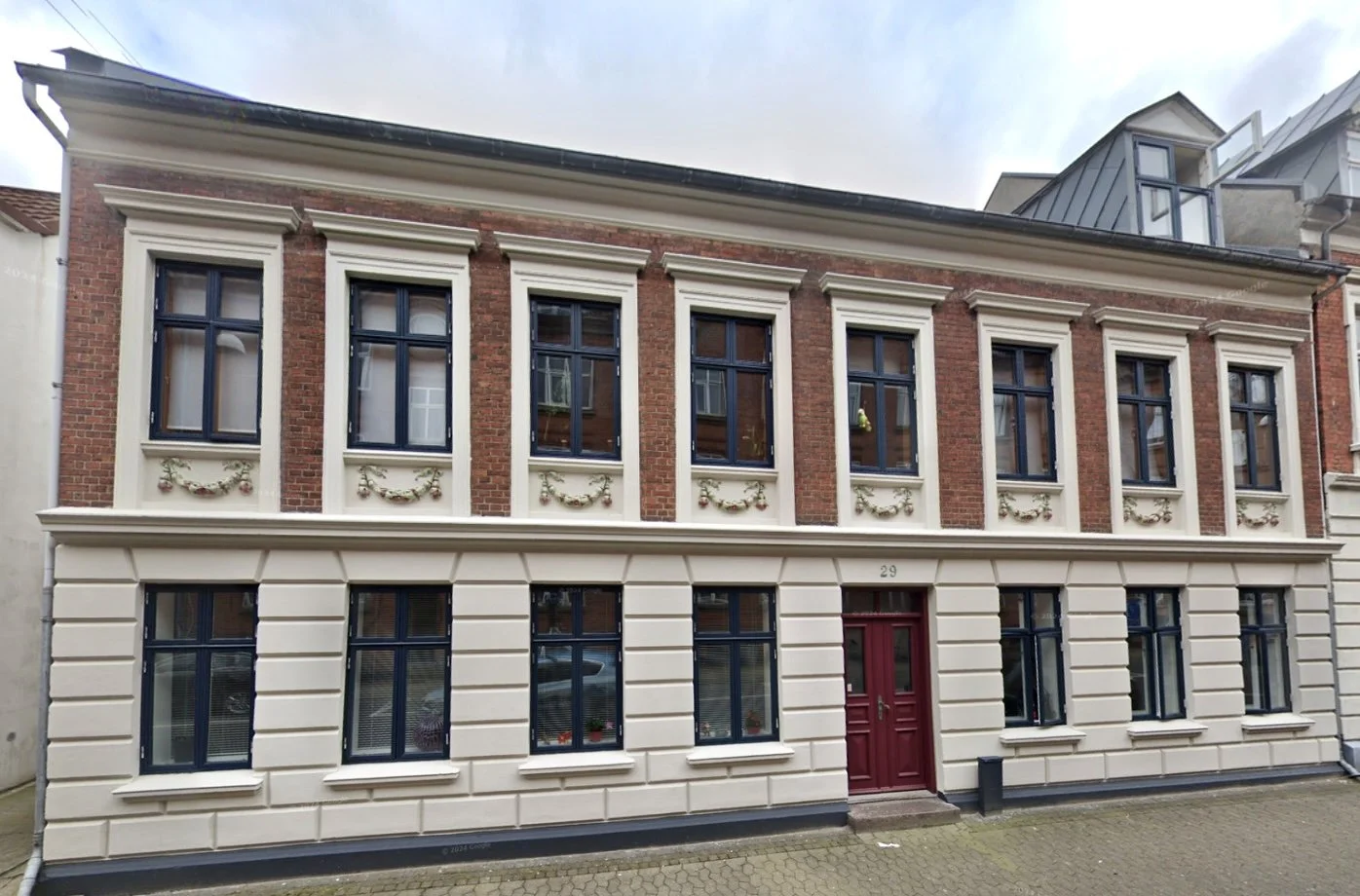 Facade af en tre-etagers murstensbygning med hvide detaljer omkring vinduer og en rød dør.