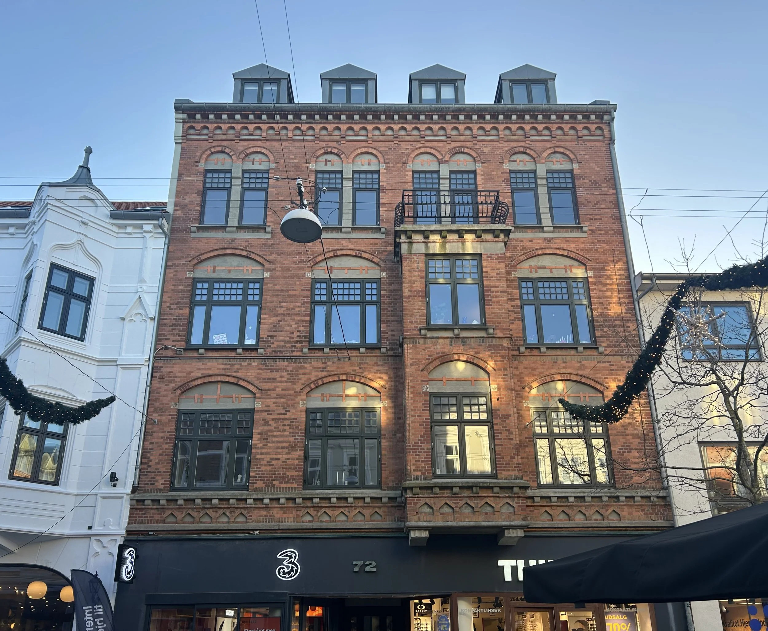 Billedet viser en bygning med butikker i stueetagen, herunder en isbutik med pink skilt og en tøjbutik, med en overbygning af murstensbygning. Der er også nybyggeri under renovering til højre med gitter og vægge dækket af lærred.