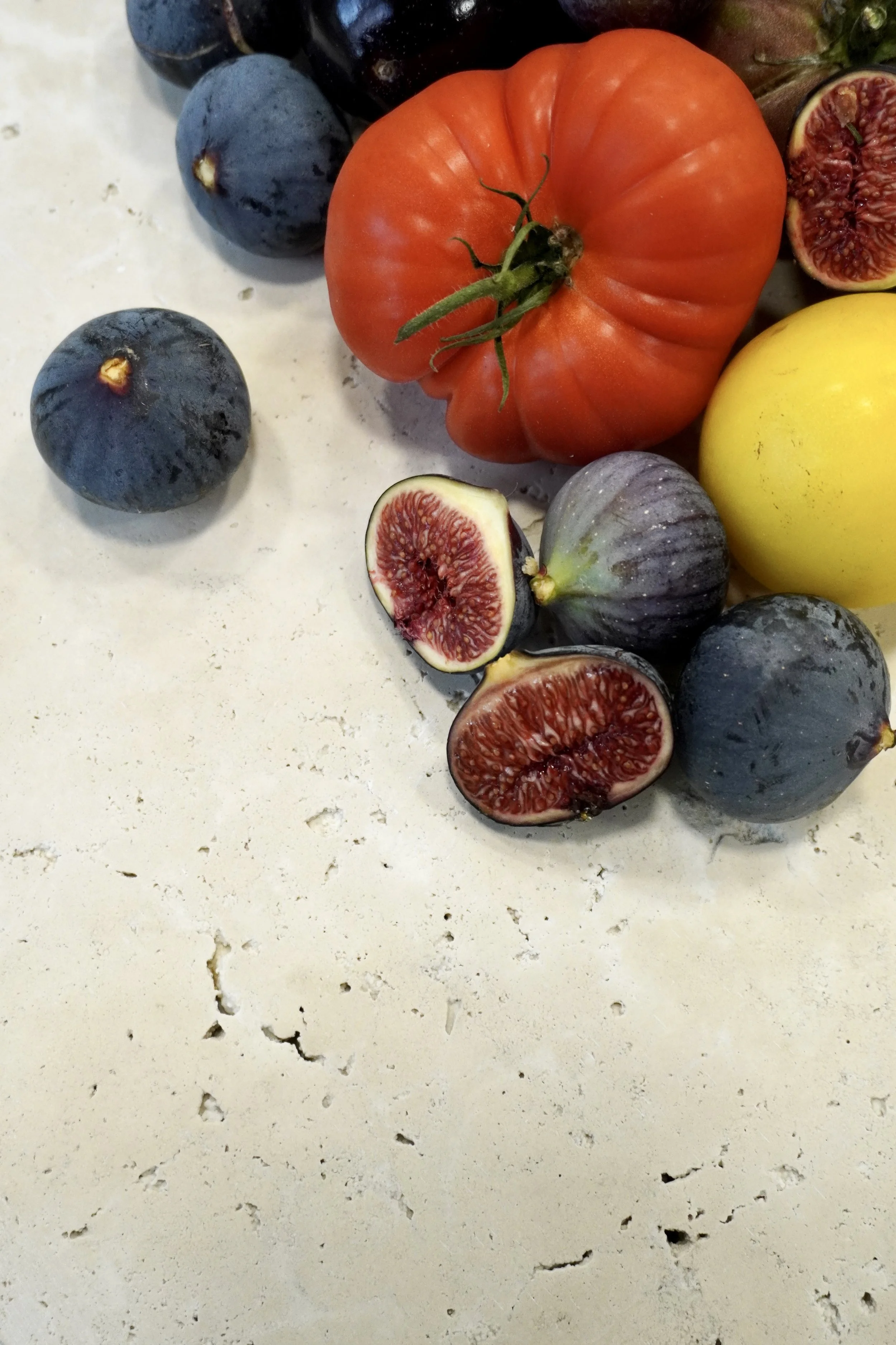 Un assortiment de fruits, comprenant des figues coupées, des tomates, des raisins et un fruit jaune, sur une surface claire.