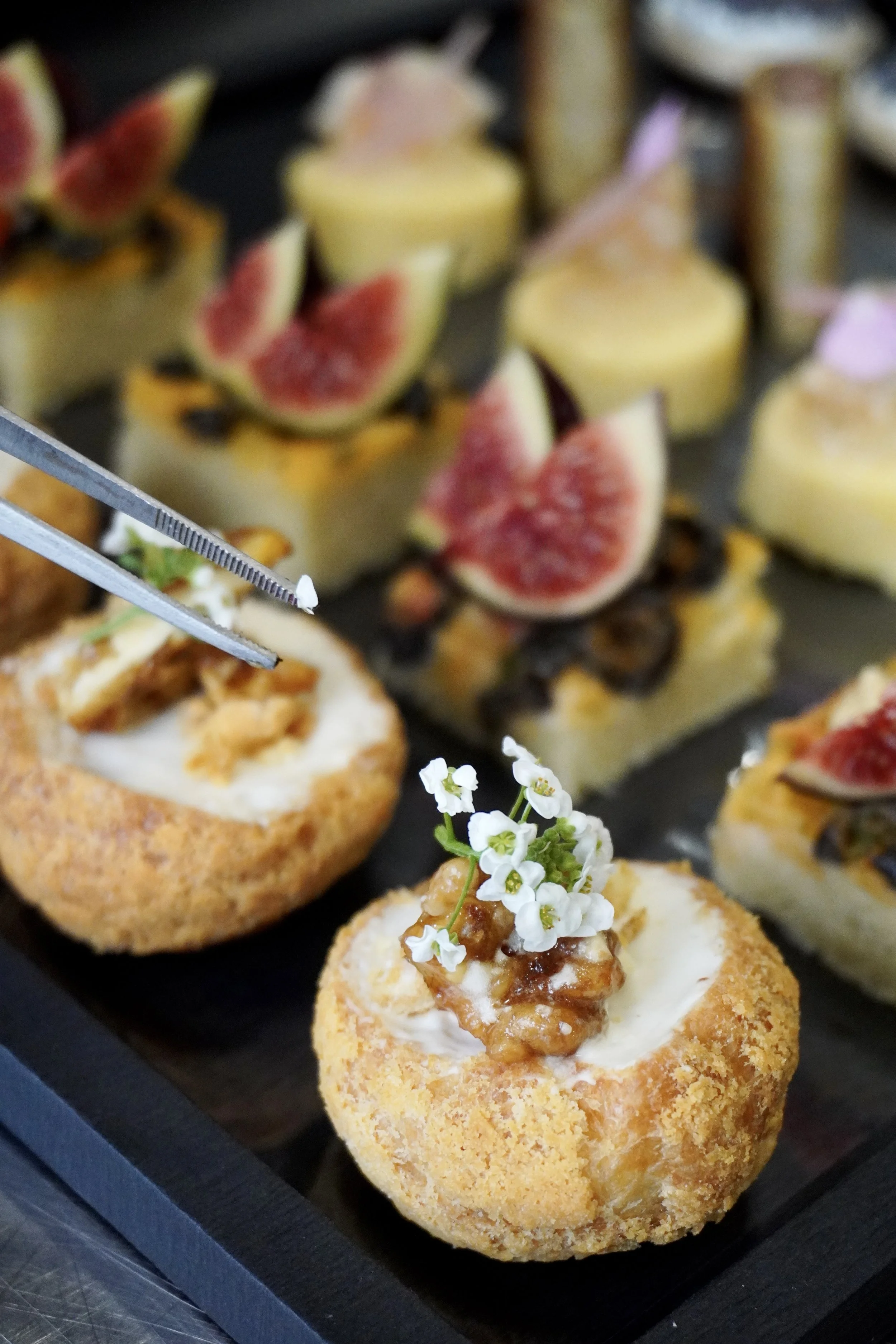 Vue rapprochée de petits amuse-bouches avec des figues, des garnitures crémeuses et des fleurs comestibles, présentés sur un plateau noir.