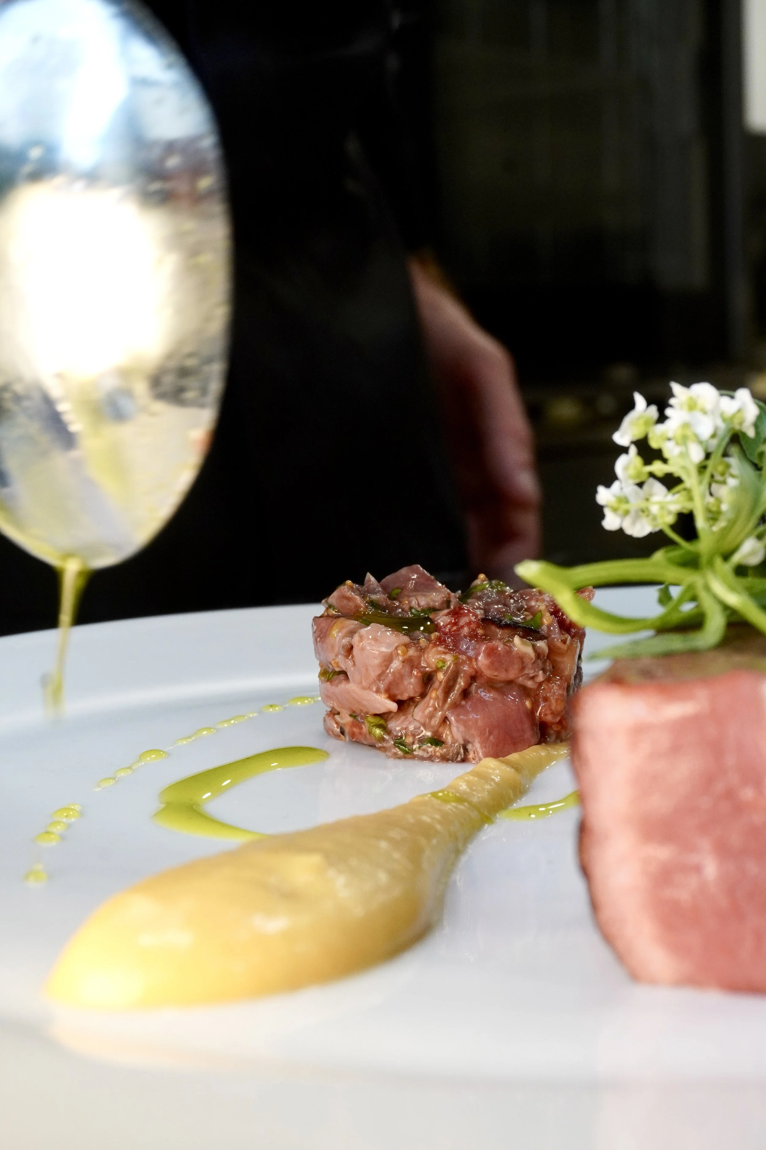 Assiette gastronomique avec un morceau de viande, une sauce, une fleur comestible, un cube de viande ou de pâté, et des gouttes d'huile ou de sauce verte, avec un verre en arrière-plan.