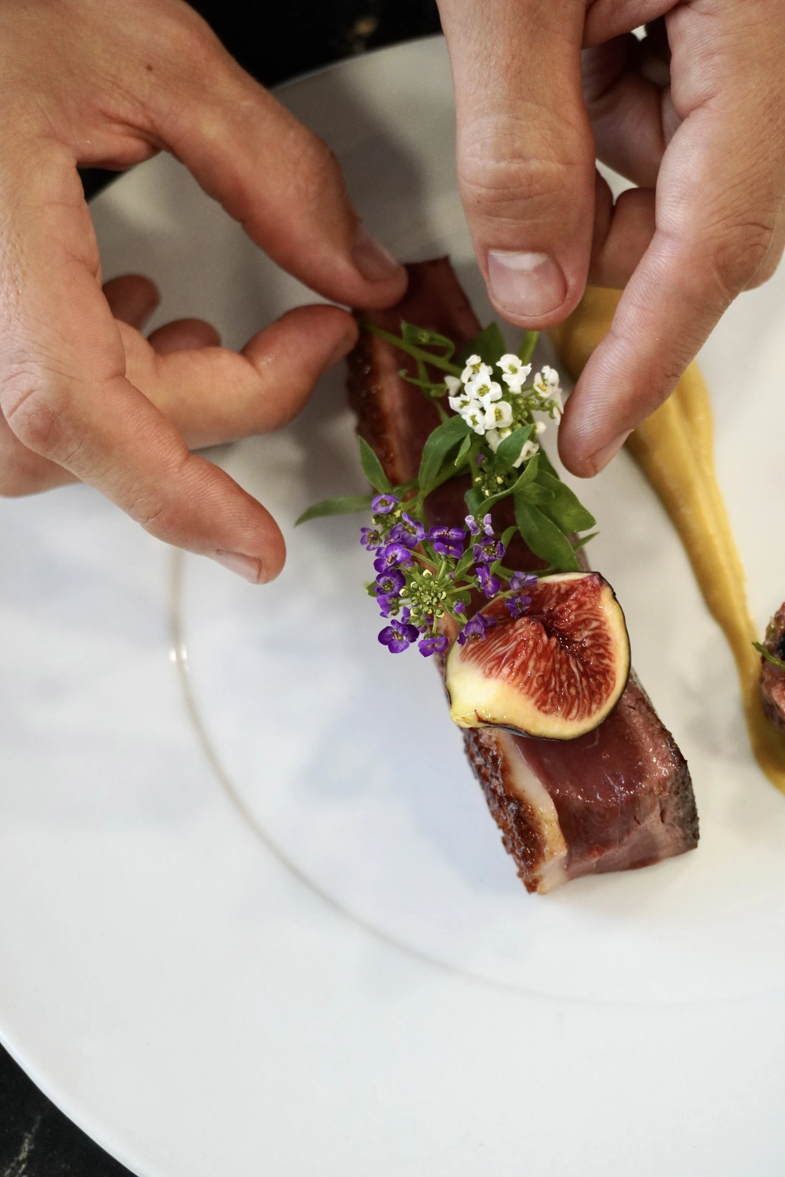 Une assiette gastronomique avec une tranche de viande, décorée avec des fleurs et un figue, accompagnée de deux sauces dans des tubes.

