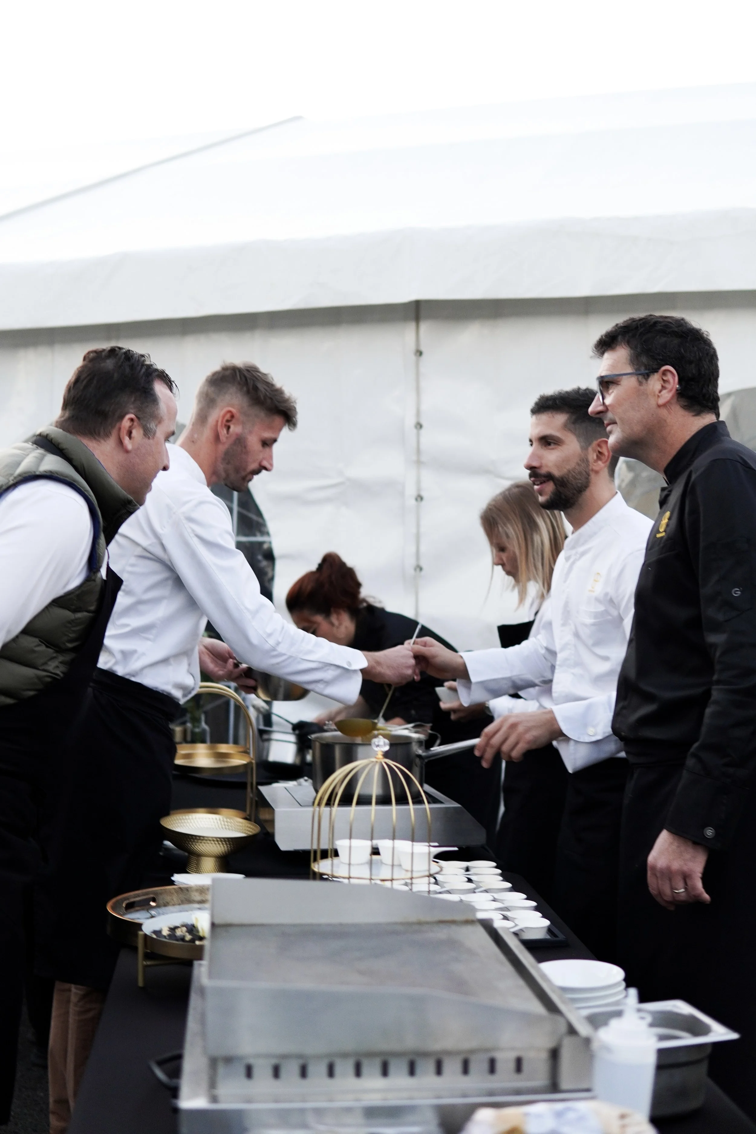 Cuisiniers en train de servir des plats lors d'un événement culinaire, avec un stand de cuisine en arrière-plan.