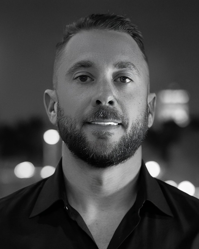 Black and white portrait of a man with short hair, a beard, wearing a collared shirt, standing outdoors at night with blurred city lights in the background.