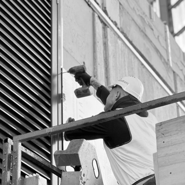 Construction worker installing outdoor air conditioning unit on building exterior, wearing safety helmet and working from scaffold.