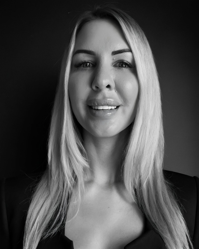 Black and white portrait of a smiling blonde woman with long hair, wearing a dark top, against a dark background.