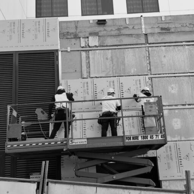 Three construction workers wearing safety helmets and gloves work on a building's exterior on a raised platform, installing or inspecting insulation panels.