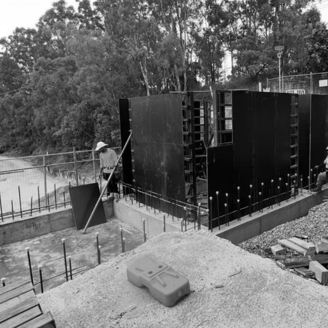 Construction site with black formwork panels being assembled, a worker wearing a helmet and safety gear, construction tools and materials scattered around, and a natural wooded area in the background.