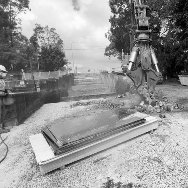 Construction site with a large excavator crushing metal sheets into smaller pieces, worker in helmet and safety gear watching nearby.
