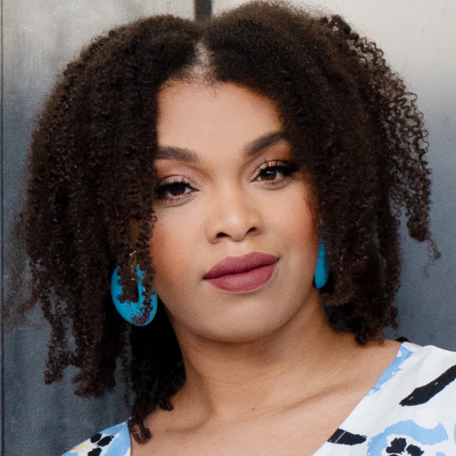 Close-up of a woman with curly hair, wearing blue earrings and a patterned top, looking confidently at the camera.