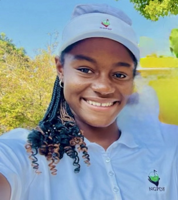 Young woman smiling outdoors, wearing a white cap and jacket, with trees and a grassy field in the background.