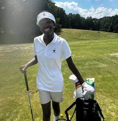 A woman smiling on a golf course, wearing a white polo shirt and shorts, holding a golf club, with a golf bag nearby, and trees and blue sky in the background.