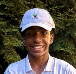 Young person wearing a white hat and shirt smiling outdoors with trees in the background.