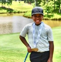 Young boy smiling on a golf course, wearing a cap, light shirt, and holding a golf club.