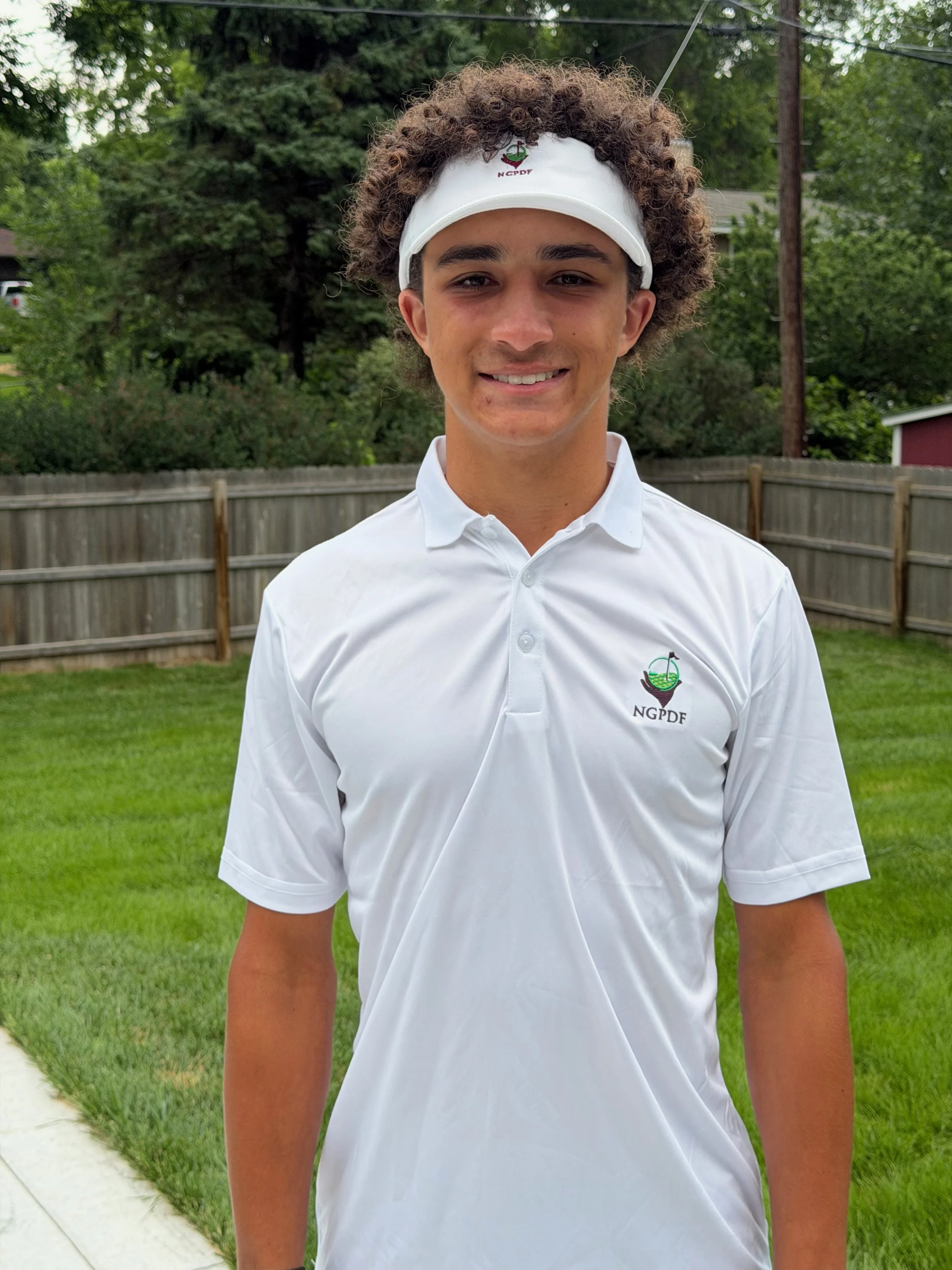 Young woman with curly hair wearing a white Titleist golf cap, a blue long-sleeve shirt, and a black puffer vest, standing on a golf course.