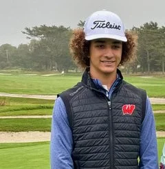 Young woman with curly hair wearing a white Titleist golf cap, a blue long-sleeve shirt, and a black puffer vest, standing on a golf course.