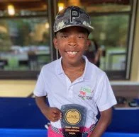 Young boy wearing a white school uniform holding a sports award plaque, smiling, with a building and trees in the background.