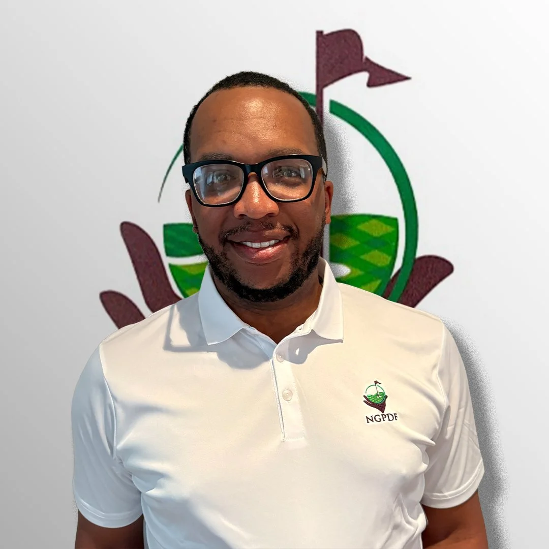 A man with glasses wearing a white polo shirt with a logo, standing in front of a wall with a logo featuring green, purple, and black elements.