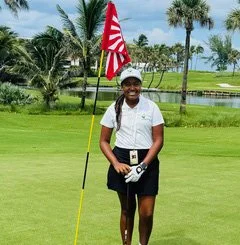 A woman standing on a golf course next to a flagstick with an American flag, holding a golf club, with palm trees and water in the background.
