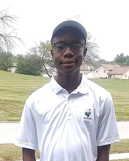 Young man wearing sunglasses, a blue sports jacket, and a medal around his neck, smiling inside a building with large windows and a view of trees outside.