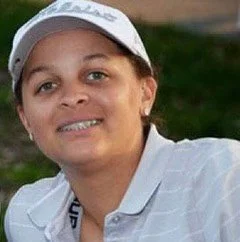 Close-up of a woman wearing a white cap and polo shirt, smiling outdoors with a grassy background.