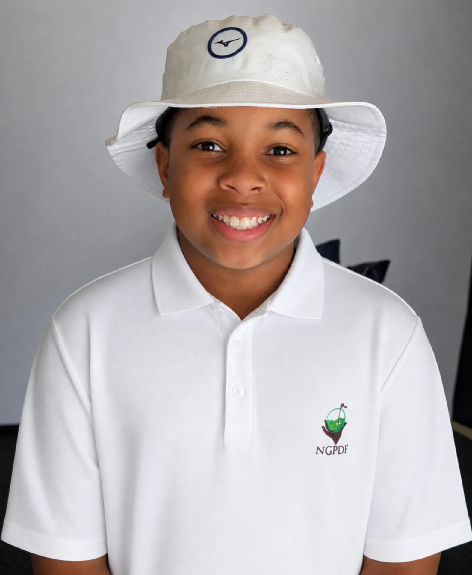 Young boy smiling on a golf course, wearing a cap, light shirt, and holding a golf club.