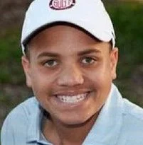 Close-up of a smiling young boy wearing a white baseball cap and a light blue shirt outdoors.