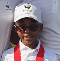 Young boy wearing sunglasses, white cap with green logo, white shirt, and red lanyard, smiling outdoors.