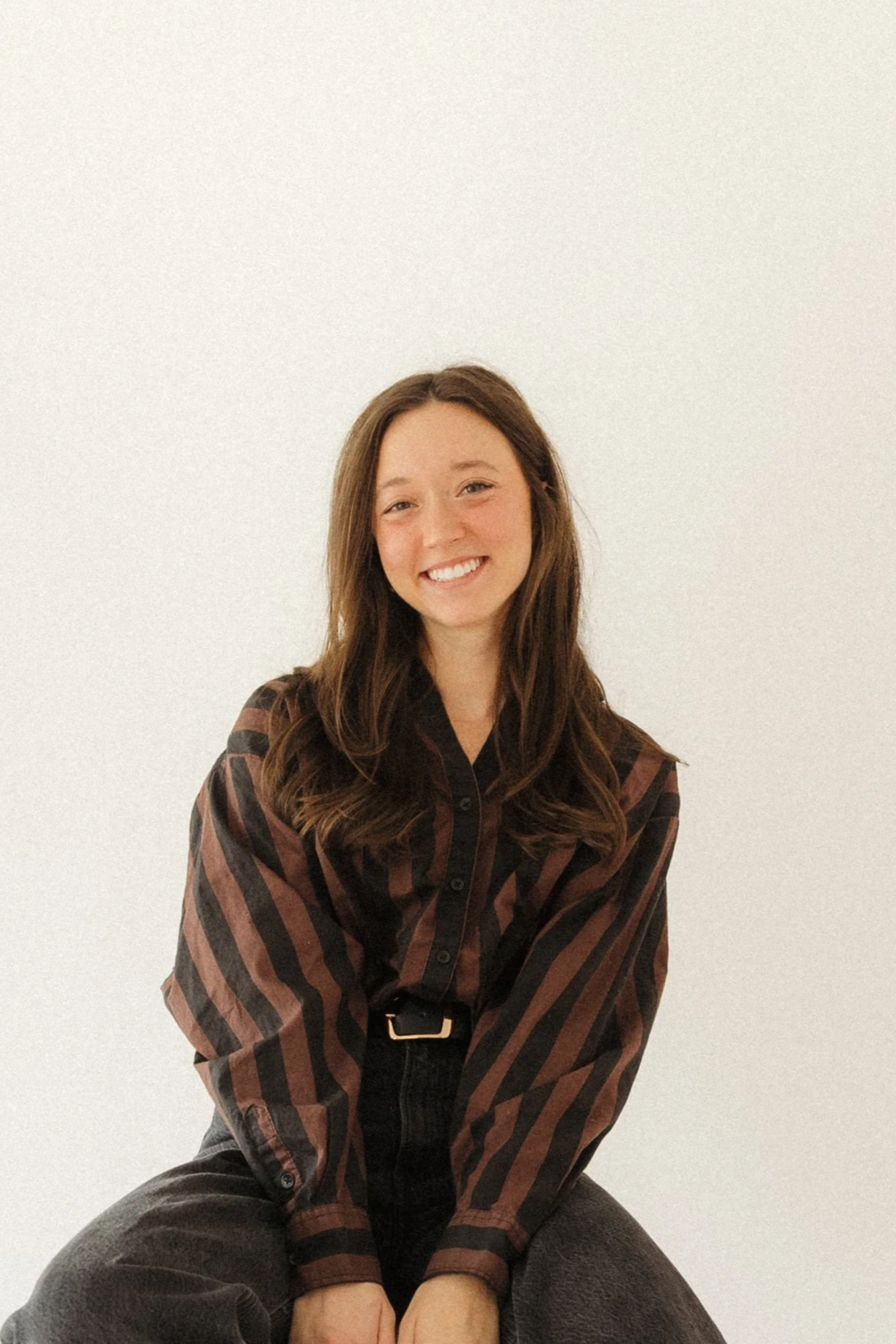 A young woman with wavy brown hair smiling while sitting on a brown armchair in a cozy room, with a green plant in the background and warm lighting.