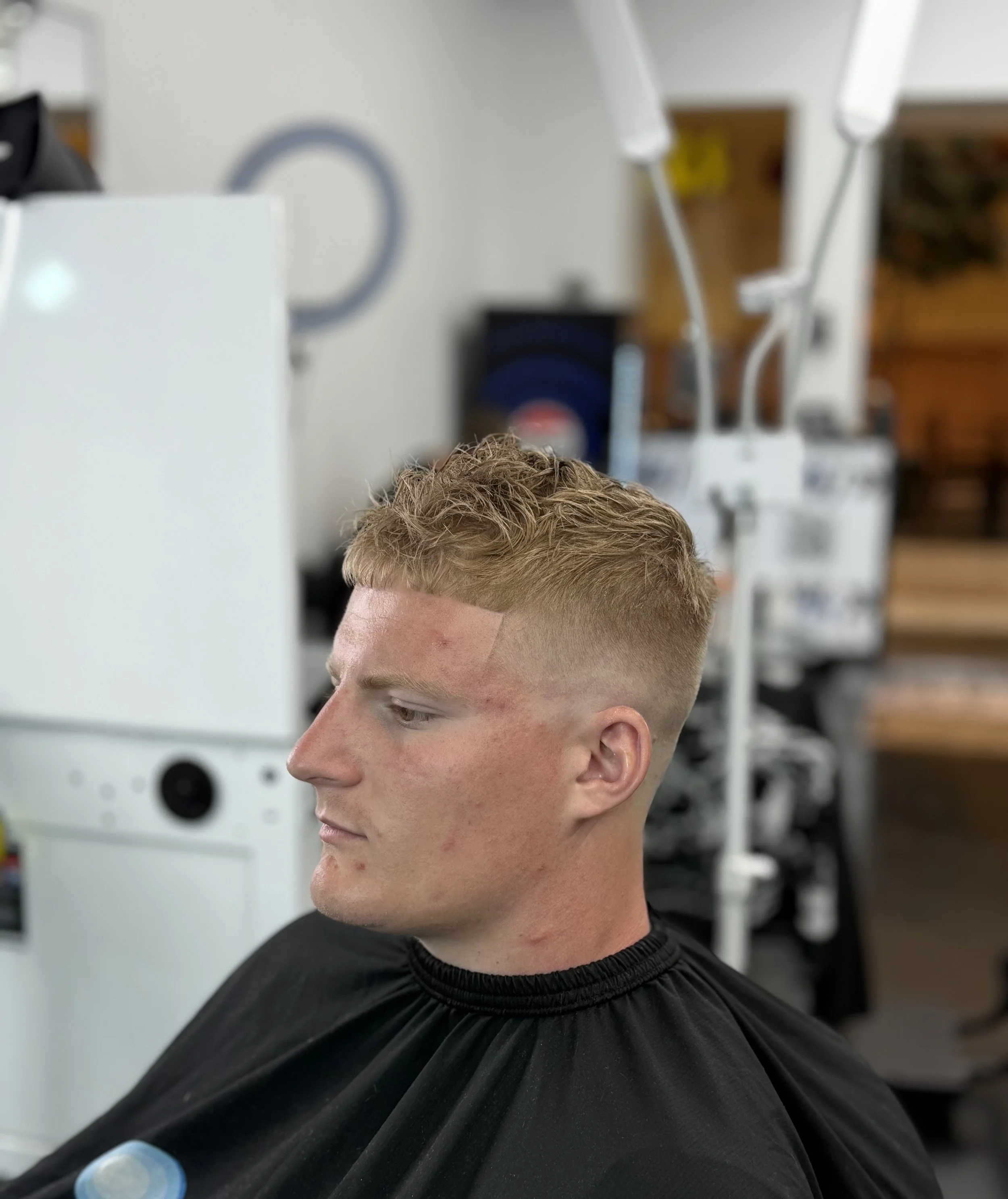 Young man with blonde hair getting a fresh haircut in a barbershop, seated with a black cape around his shoulders, in front of barber equipment.