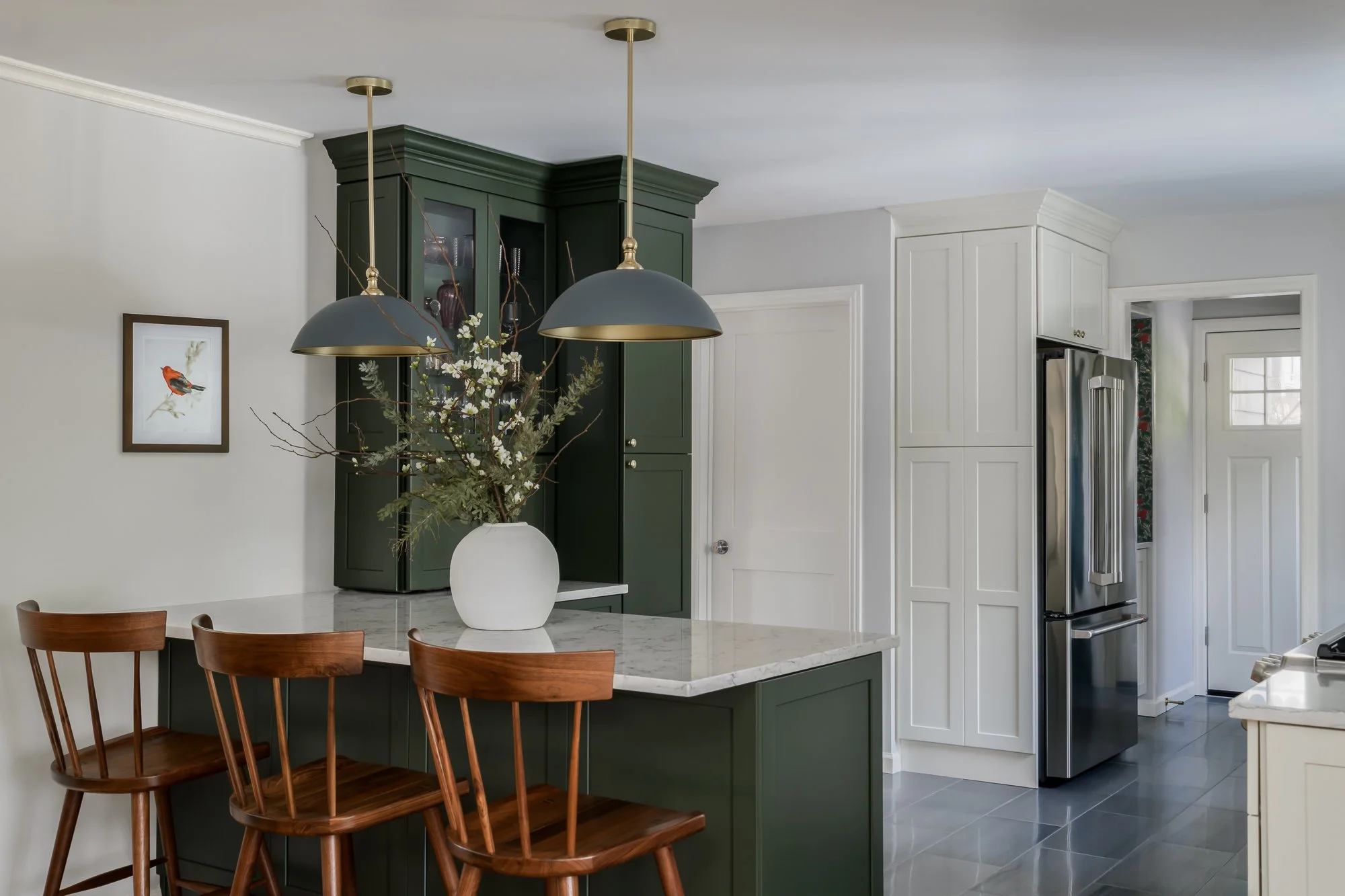 Kitchen with green cabinets, white walls, and a marble countertop island with three wooden chairs. Two black pendant lights hang above the island, and a white vase with branches and flowers is on the counter. A picture of a bird hangs on the wall, and stainless steel refrigerator is near a door with window panes.