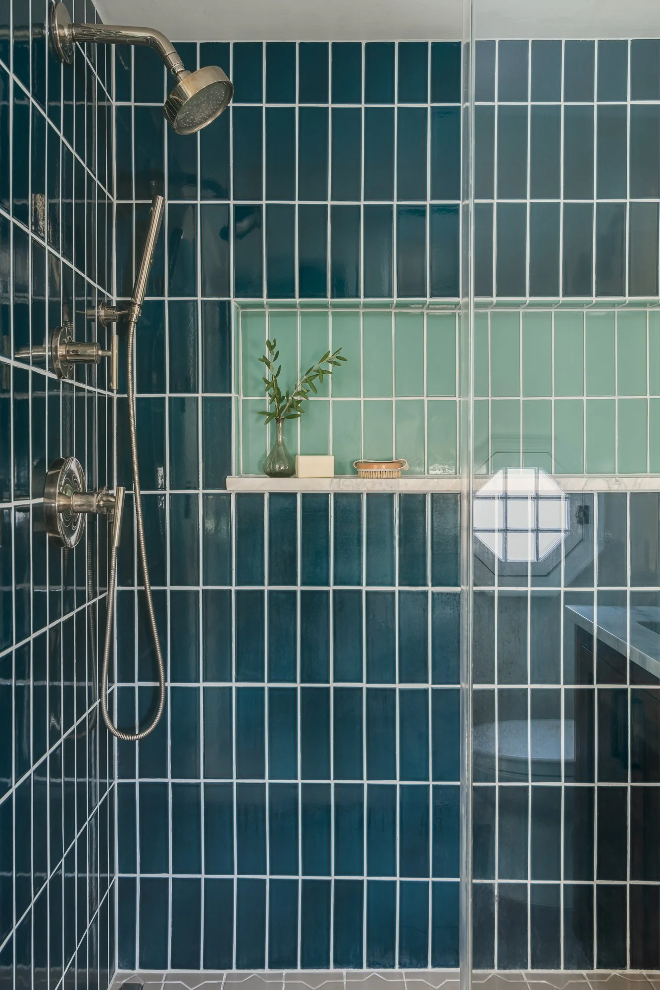 A modern shower area with dark blue tiles on the left and green tiles on the back wall, featuring polished nickel fixtures, a small shelf with a vase and toiletries, and a glass partition.