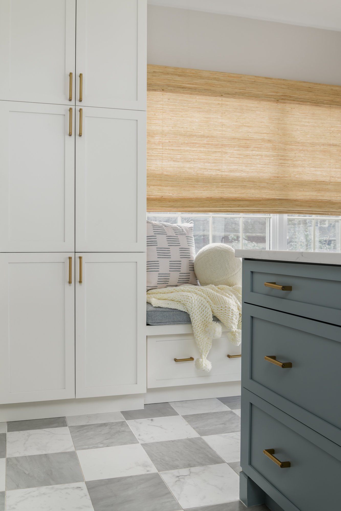 Corner of a classic kitchen with white cabinets, a blue drawer unit, and a built-in window seat with pillows and a blanket. There is a window with a bamboo shade and a blue and cream checkered pillow.
