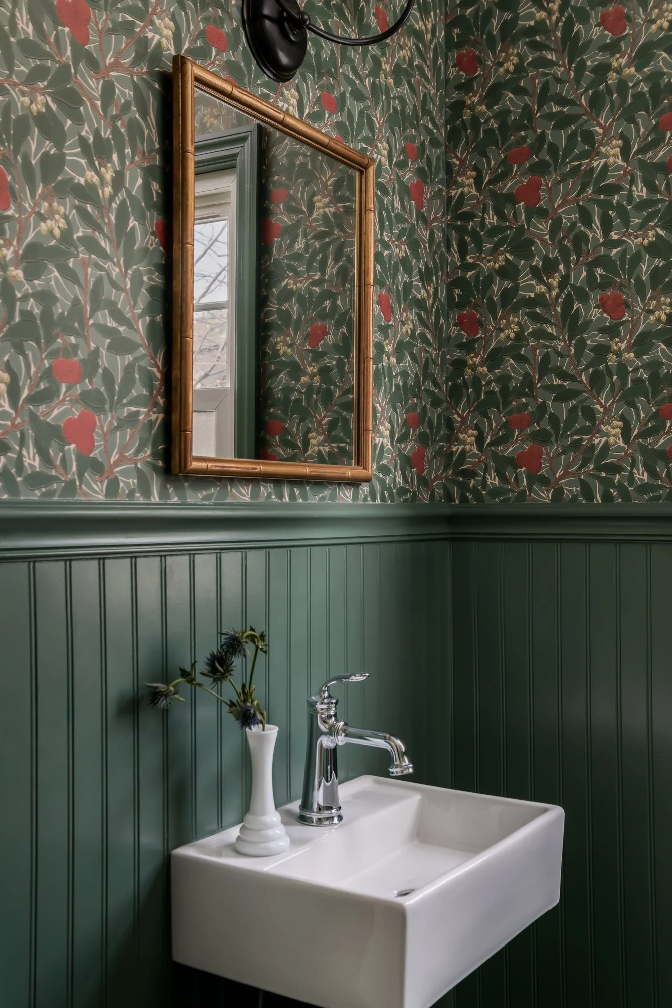 Powder room with botanical wallpaper, an antique mirror, wall sconce, a white wall-mounted sink with chrome fixtures, painted beadboard and decorative vase with flower.