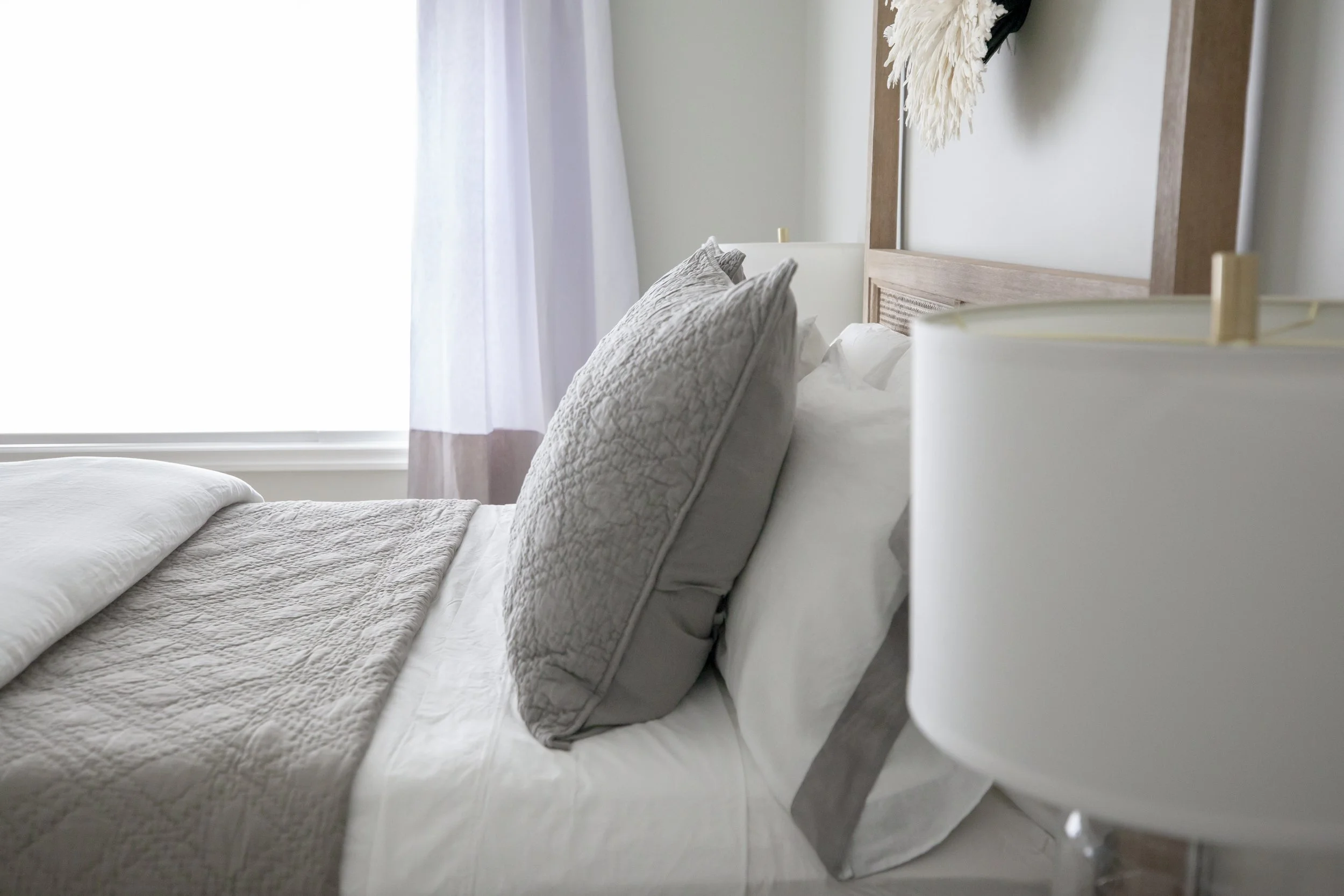 A  serene bedroom with gray pillows and bedding, next to a nightstand with a lamp, in a bright bedroom with sheer white curtains.