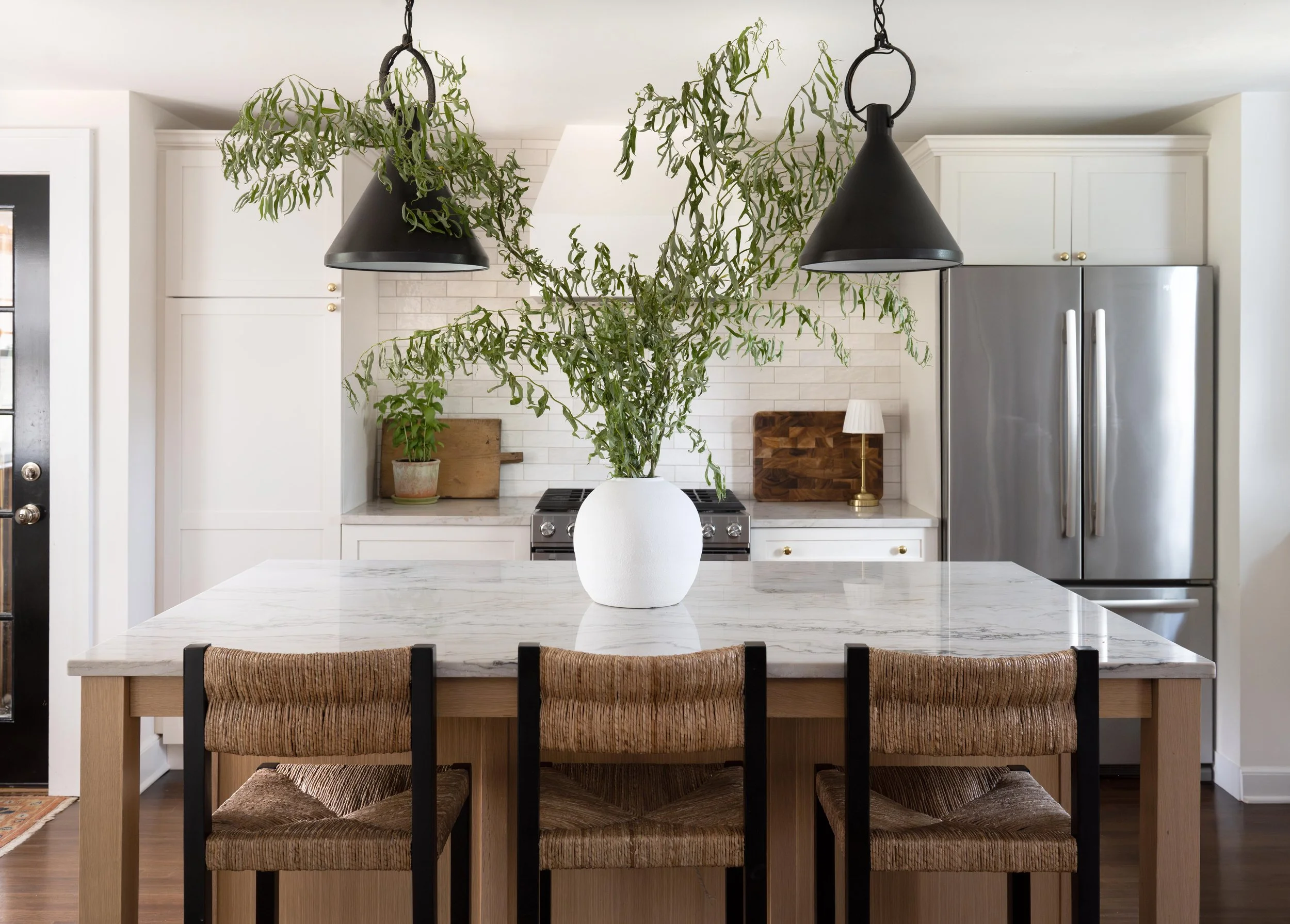 Kitchen with white cabinets, stainless steel refrigerator, marble island with wicker chairs, large vase with greenery, and black hanging lights.