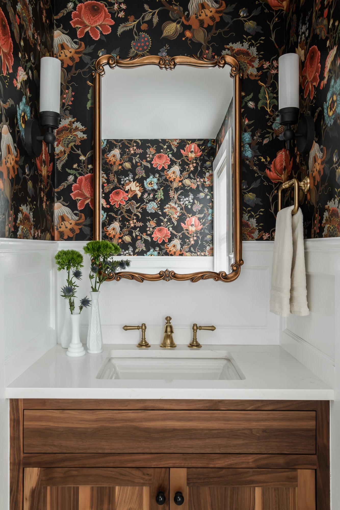 Sophisticated powder room with floral wallpaper, a gold-framed mirror, wall sconces, a white sink with brass fixtures, wooden cabinet, and decorative vases with flowers.