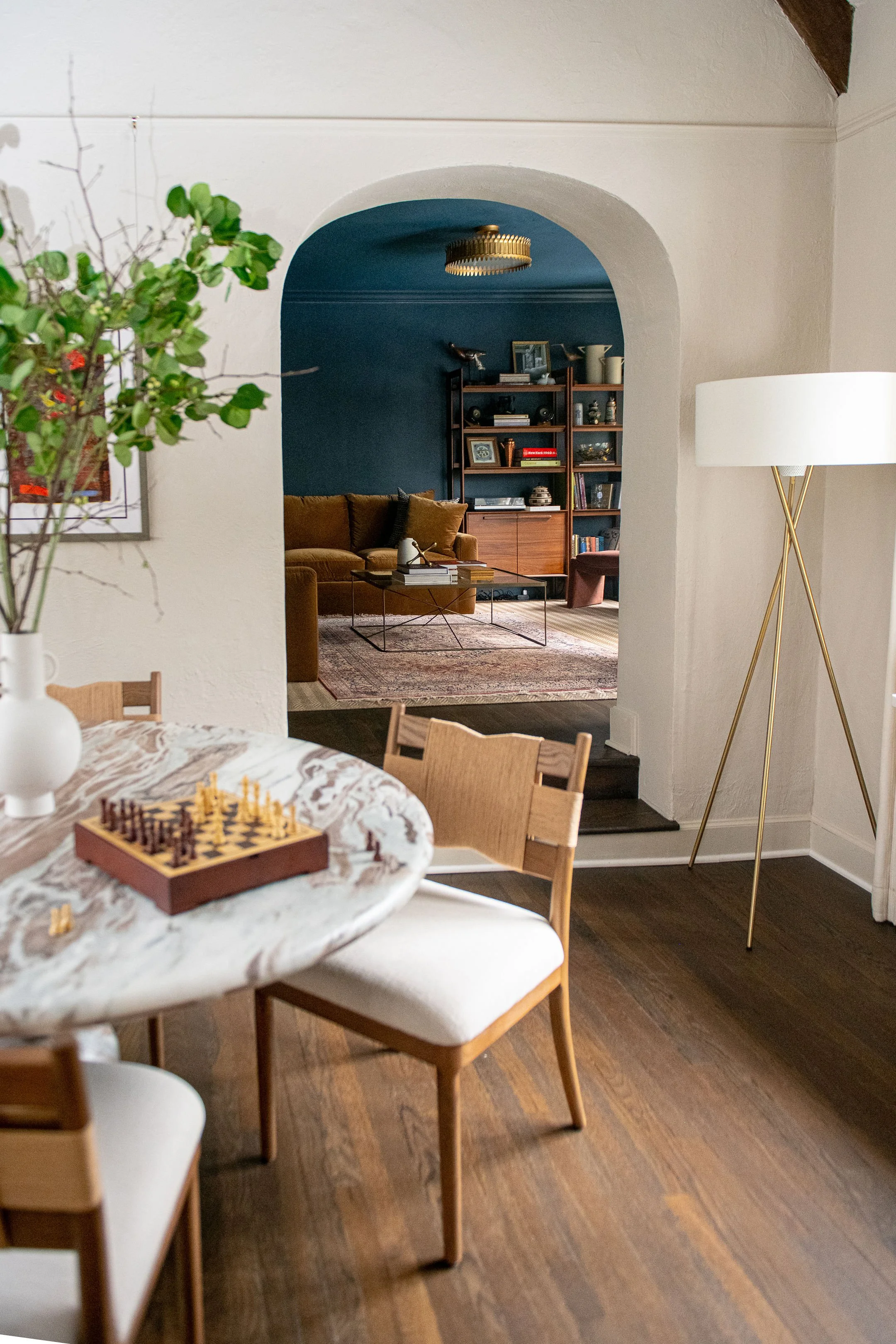 A view through an arched doorway of a living room with a blue accent wall, a brown sectional sofa with pillows, a bookshelf, and a vintage-style sideboard, with a rug on the floor.