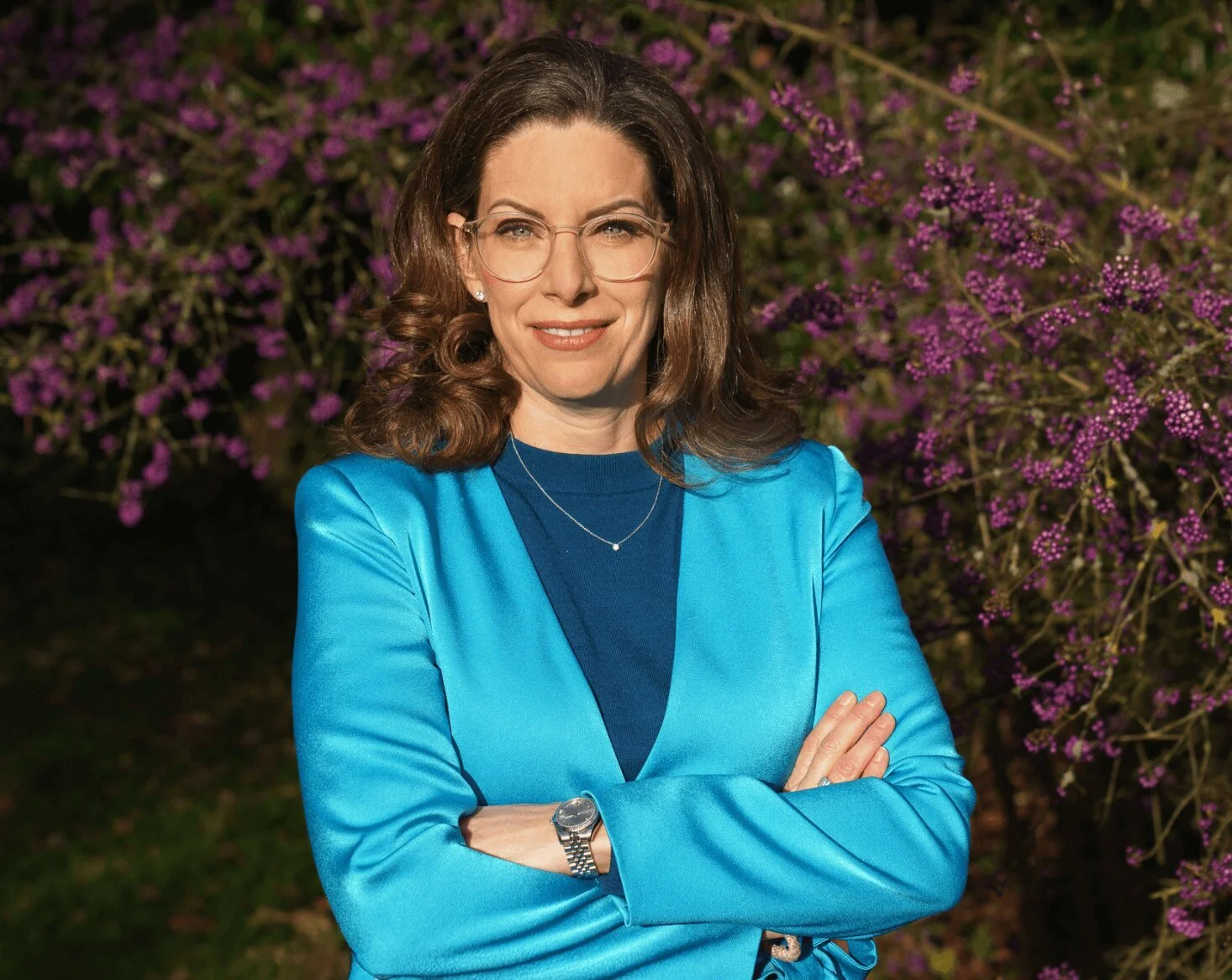 Eine Frau mit braunen, lockigen Haaren, trägt eine blaue Jacke und eine blaue Oberteil. Sie trägt eine Armbanduhr und eine Halskette, steht vor einer blühenden Pflanze mit lila Blüten, posiert mit verschränkten Armen, lächelt leicht und schaut in die Kamera.