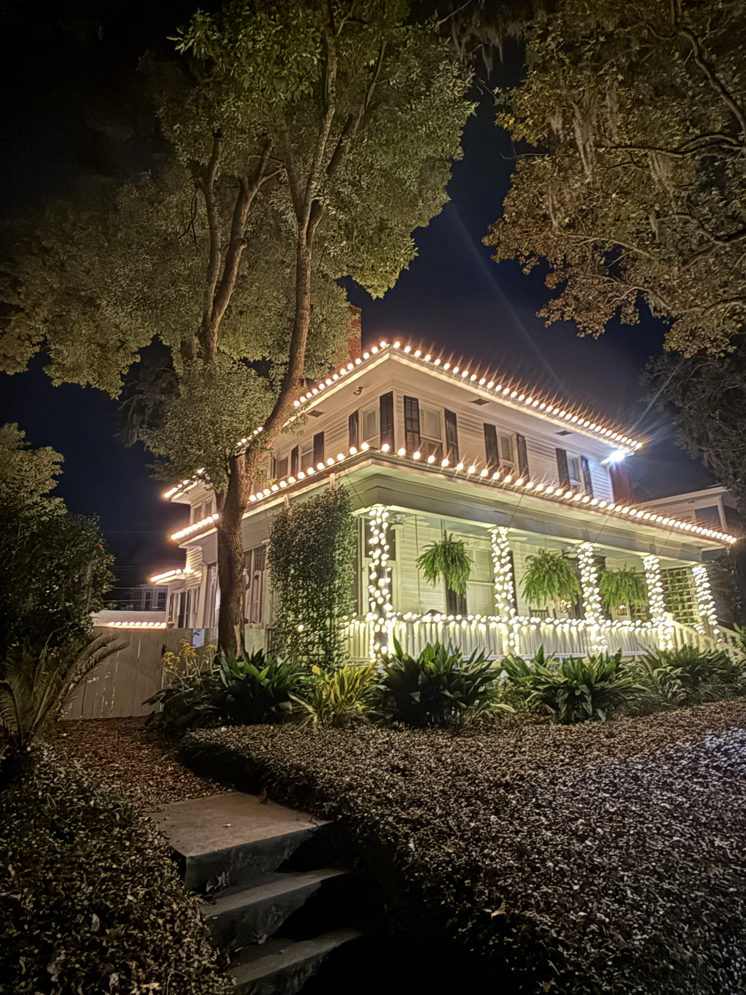 A large two-story house decorated with white string lights outlining the roof, porch, and columns, surrounded by trees and shrubs at night. savannah christmas lighting