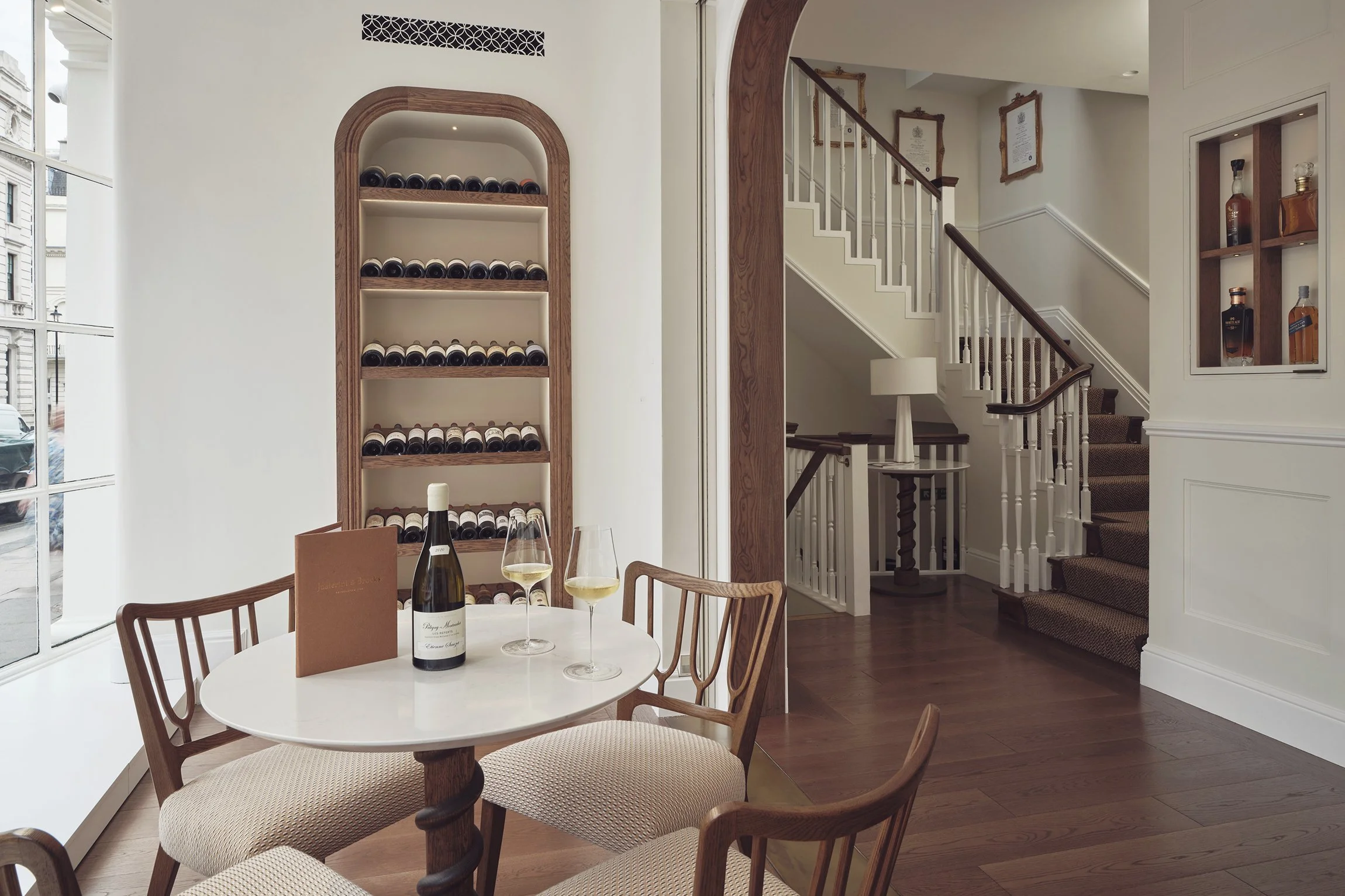 Pale room with a round marble table, antique chairs and a bespoke arched oak niche displaying wine, beside tall windows.