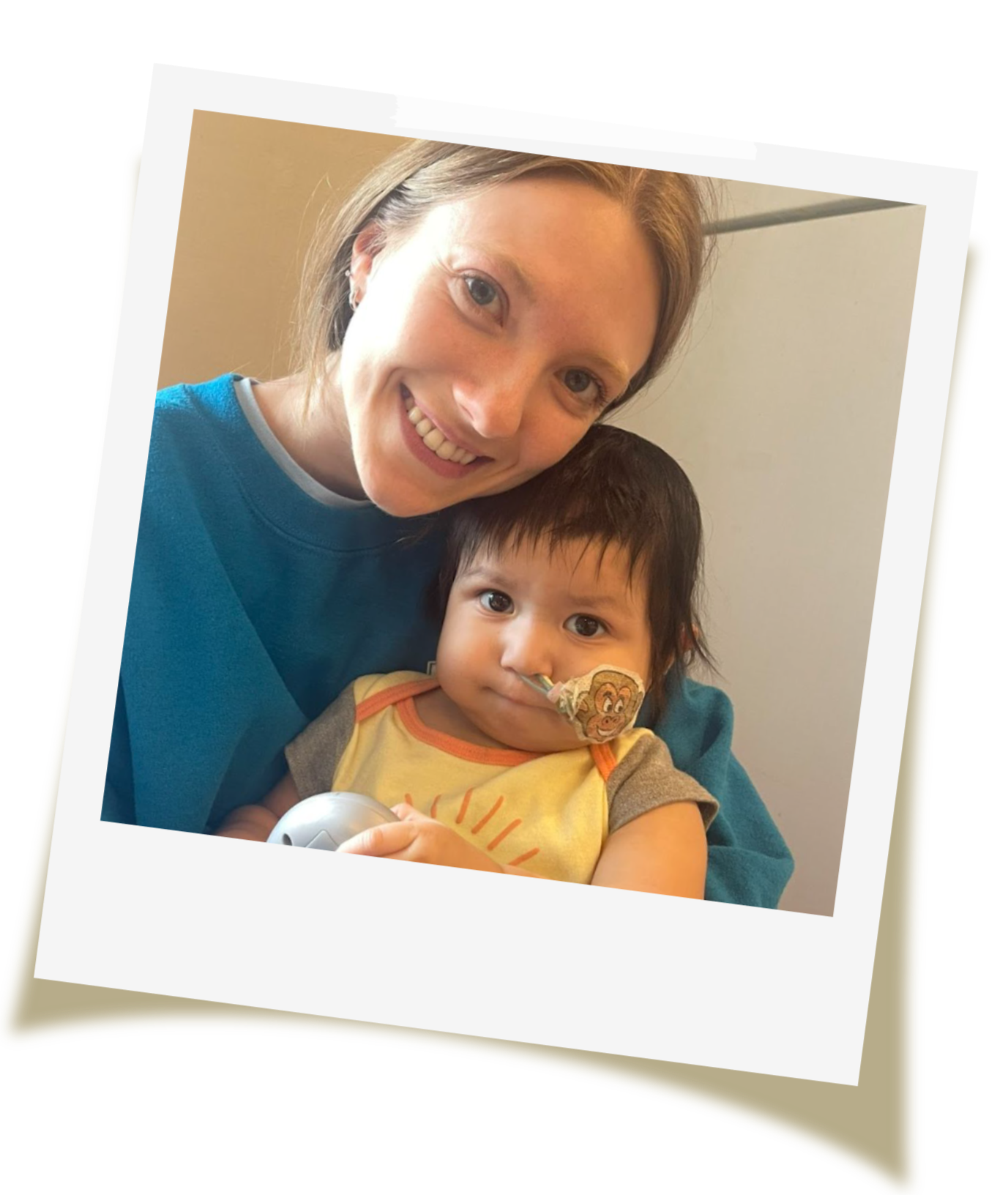 Smiling woman holding a young child with a feeding tube in his nose, indoors with a beige wall in the background.