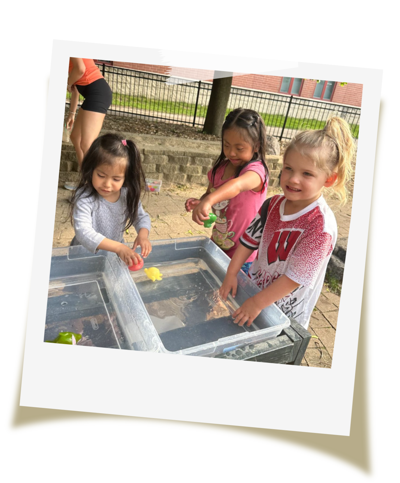 Three young girls are playing with floating toys in a water table outdoors, with a brick building, trees, and a person in background.