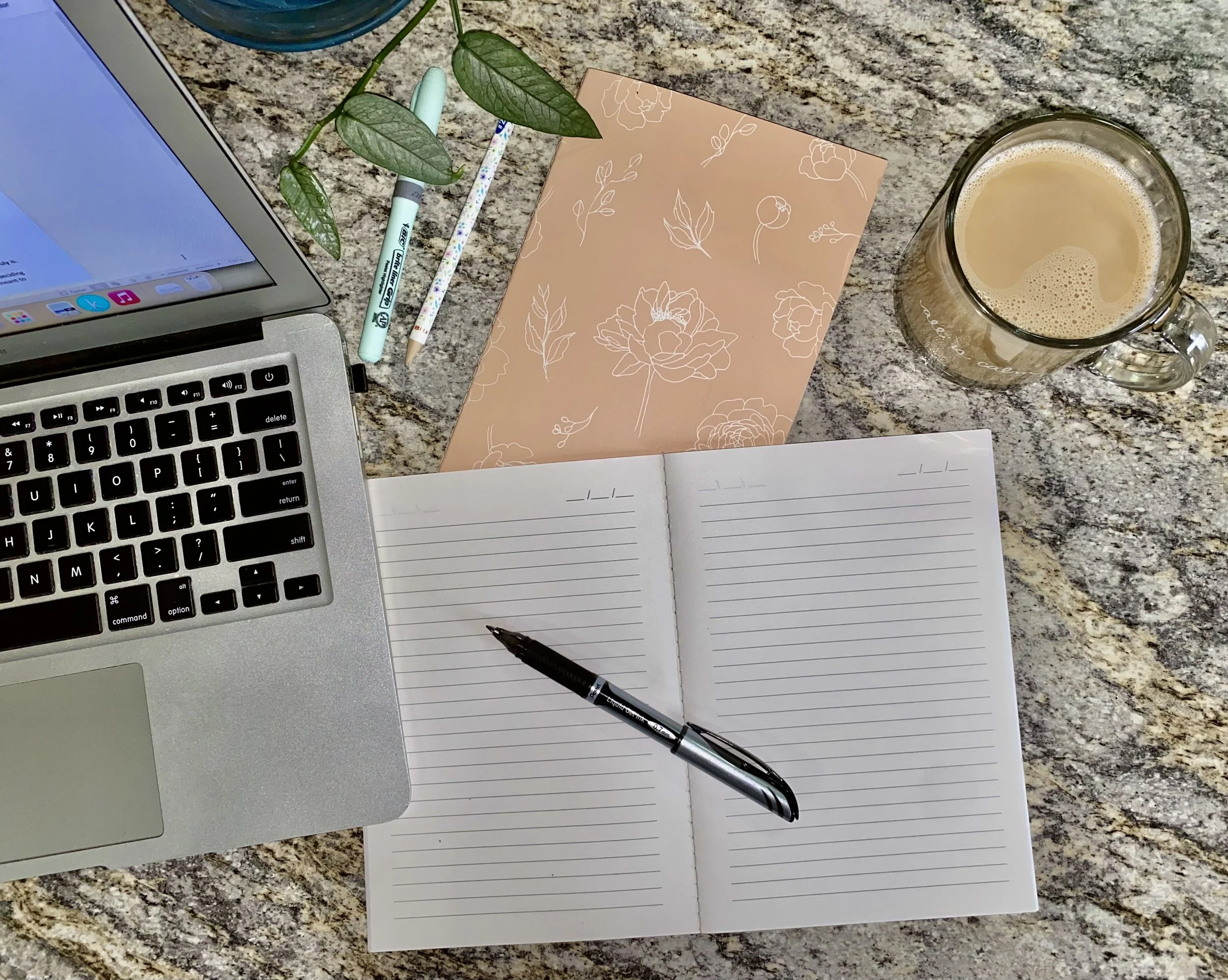 A workspace on a granite countertop with a laptop, open notebook with a pen, floral notecard, two pencils, a mug of coffee, and a small green plant.