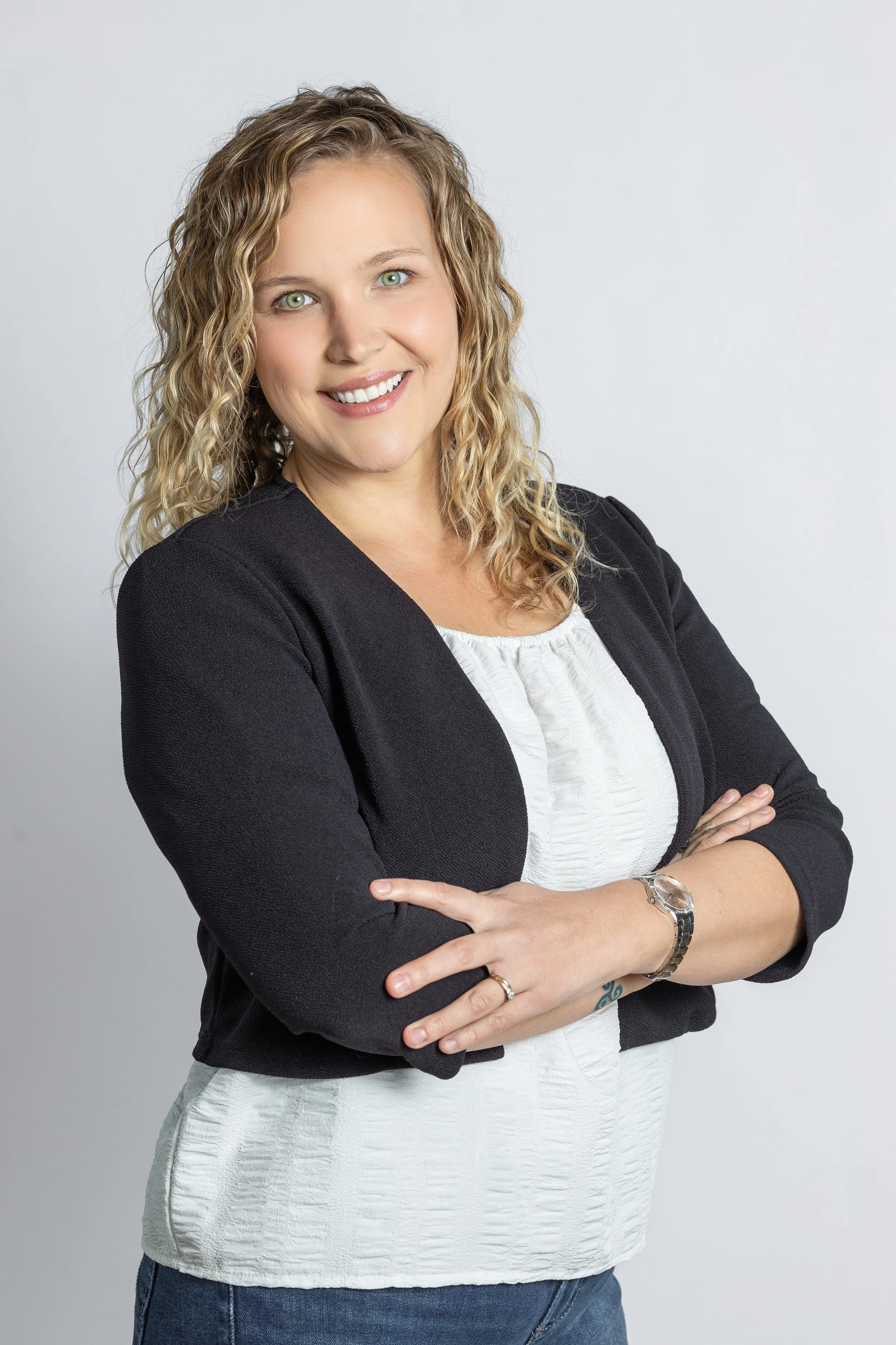 A woman with curly blonde hair smiling, wearing a black blazer over a white blouse, with her arms crossed, standing against a gray background.