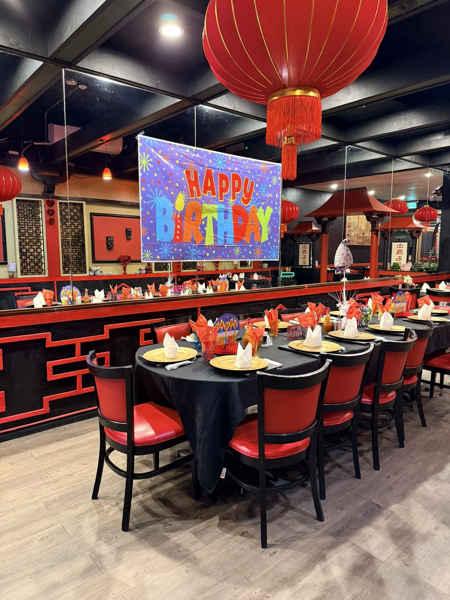 A restaurant decorated for a birthday celebration with red and gold Chinese lanterns, a large colorful 'Happy Birthday' sign, and tables set with black tablecloths, red napkins, and white plates.