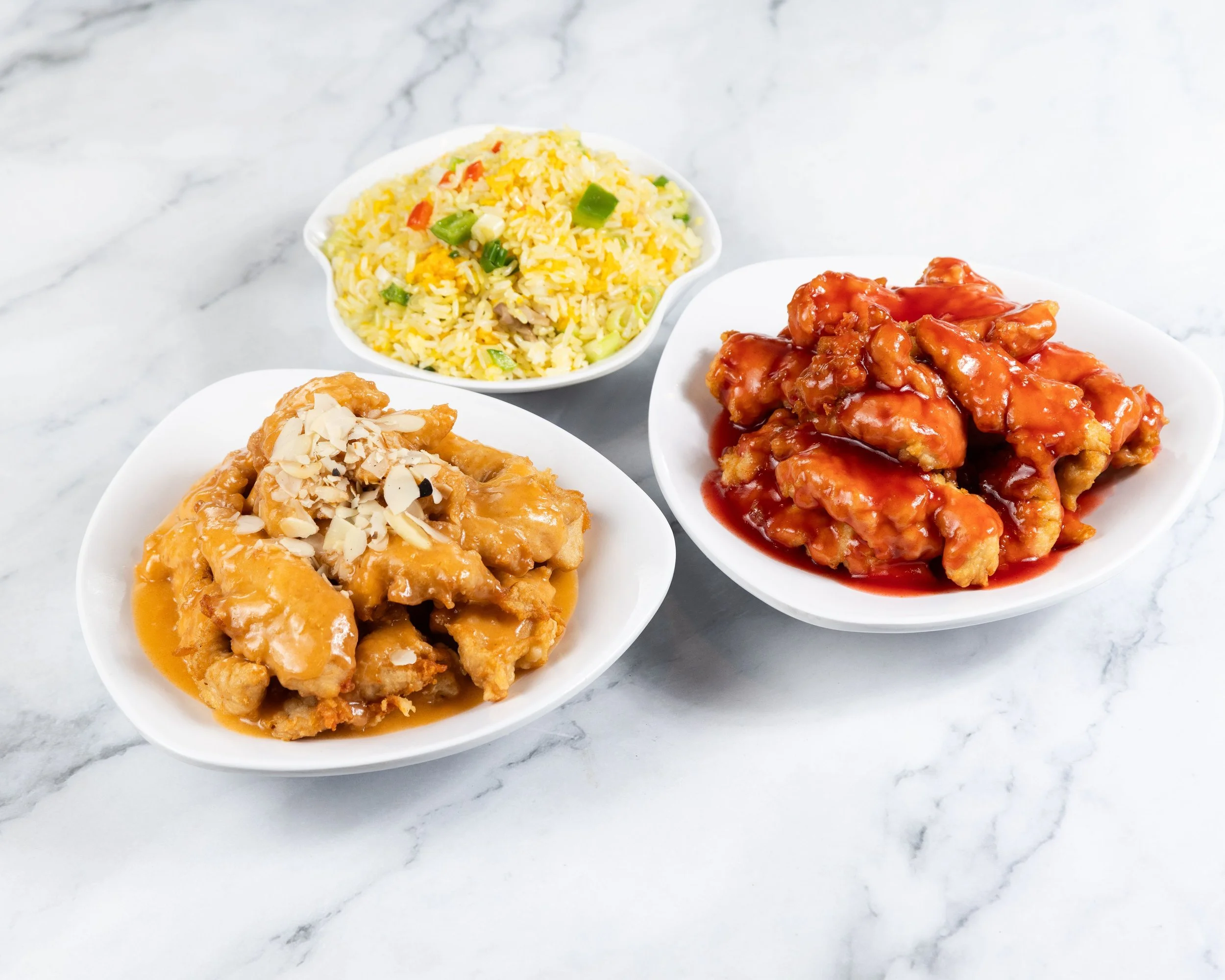 Three white plates on a marble surface, one with fried rice with vegetables, another with honey chicken topped with chopped almonds, and the third with sweet and sour chicken in red sauce.
