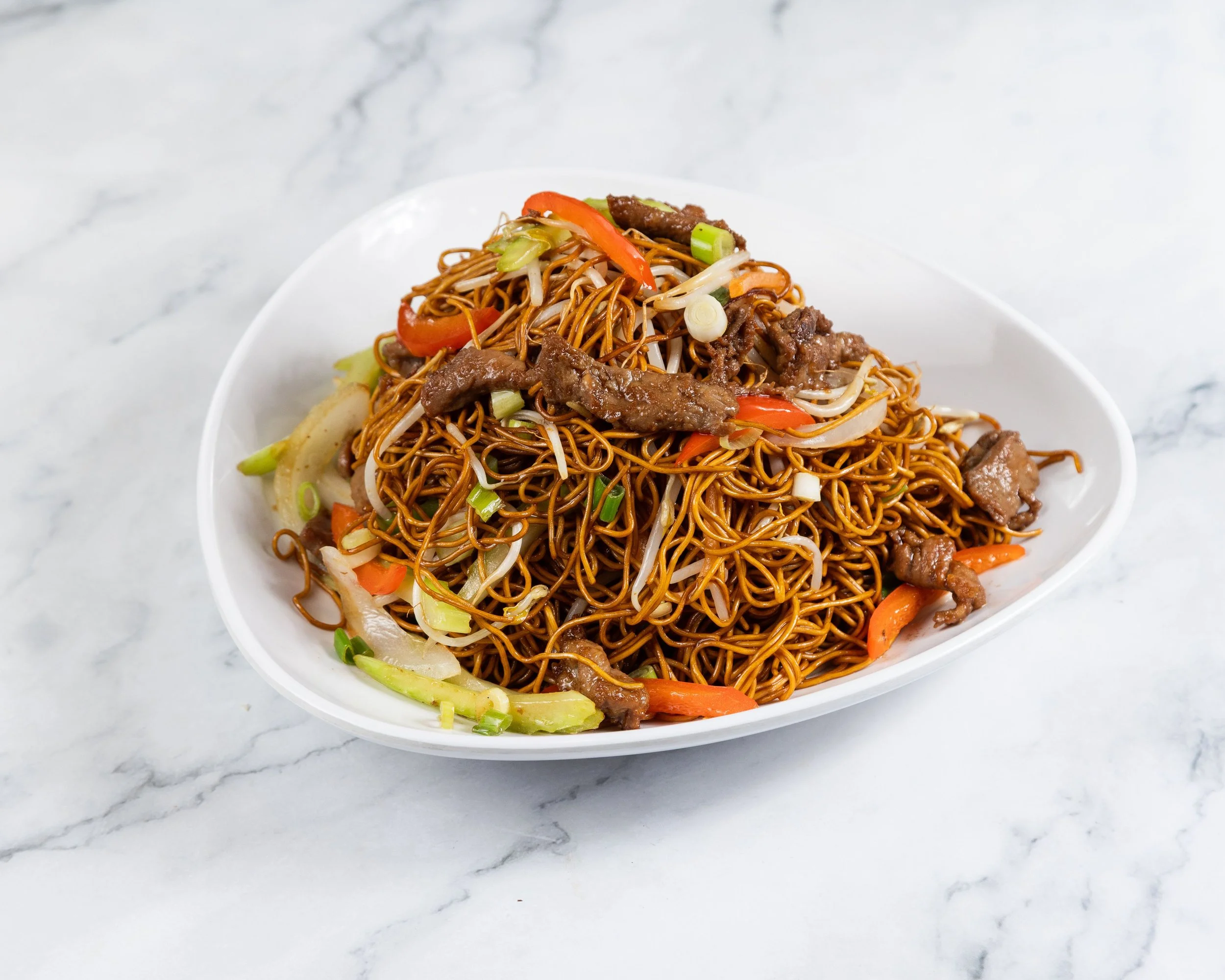 A white bowl filled with stir-fried noodles, sliced beef, and mixed vegetables such as bell peppers, onions, and green onions, placed on a white marble surface.