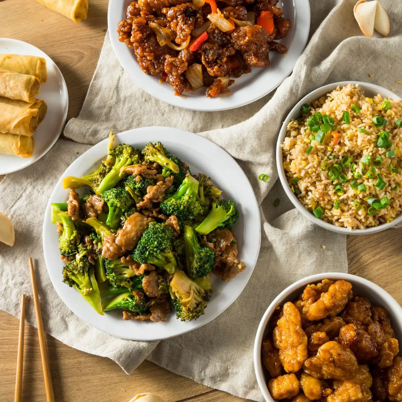 A variety of Chinese dishes on white plates: crispy egg rolls, stir-fried broccoli with beef, fried rice with green onions, sweet and sour chicken, and sweet and sour pork, arranged on a beige cloth on a wooden table.
